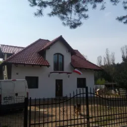 Biały dom z czerwoną dachówką, widoczny zza czarnego metalowego ogrodzenia. Na elewacji flaga Polski. Częściowo widoczny biały bus i drzewa w tle.