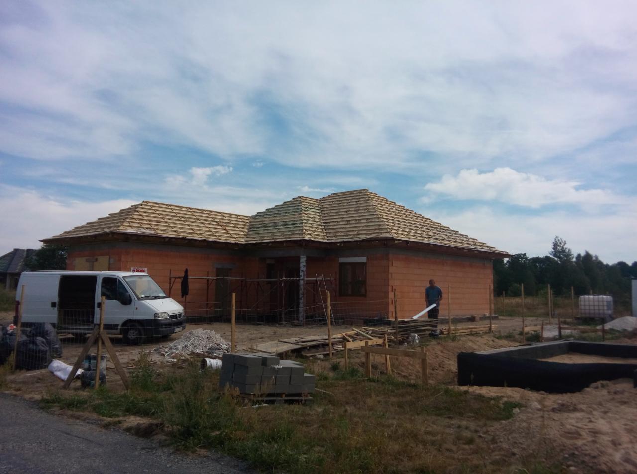 Budowa domu jednorodzinnego w Zaborze: ściany z cegły, częściowo położony dach z drewnianej dachówki, fundamenty i biały bus na posesji.
