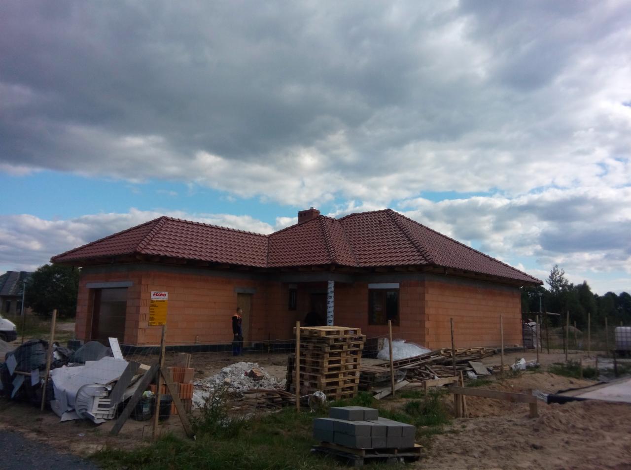 Budowa domu jednorodzinnego z czerwonej cegły, z częściowo położonym dachem z czerwonej dachówki. Widoczne palety z materiałami budowlanymi i osoba na budowie.