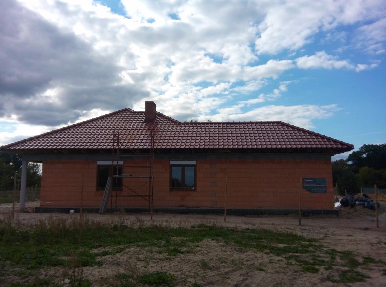Niedokończony murowany dom z czerwoną dachówką i kominem, widoczny z zewnątrz. Na pierwszym planie trawa i rusztowanie. Błękitne niebo z chmurami w tle.