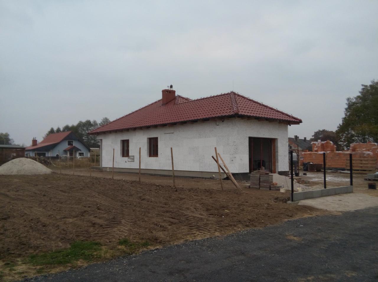 Niedokończony dom z czerwoną dachówką i widocznym ociepleniem ścian, teren przygotowany pod ogród, widok na inny budynek w tle.