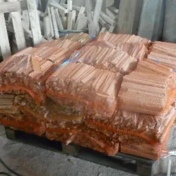 Paleta z workami wypełnionymi pociętym drewnem opałowym w siatkach, zabezpieczona niebieskim sznurkiem, składowana w magazynie z drewnianymi elementami konstrukcyjnymi w tle.