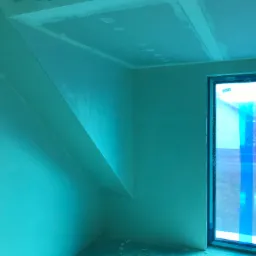 Pokój w trakcie remontu: ściany pokryte świeżą warstwą gładzi, sufit z widocznymi punktowymi łatami, okno zabezpieczone folią ochronną.