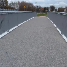 Kładka dla pieszych z szarymi barierkami po obu stronach i szarą nawierzchnią z drobnego kruszywa, widoczny znak drogowy ostrzegawczy dla dzieci w oddali.