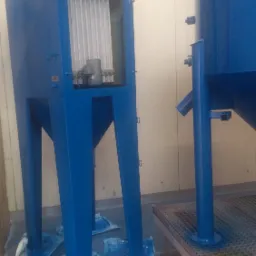 Niebieska kabina do obróbki strumieniowo-ściernej z widocznym filtrem wewnątrz, obok duży zbiornik, całość zabezpieczona folią malarską.