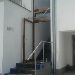 Wejście do budynku z brązowymi drzwiami zewnętrznymi z przeszklonym daszkiem, schodami wykończonymi niebieską taśmą malarską oraz metalową barierką.