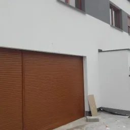 Zamknięta, brązowa brama rolowana garażu w nowoczesnym domu z białymi ścianami i szarymi akcentami, widoczne okna na piętrze.