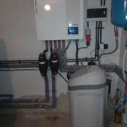 Biały piec centralnego ogrzewania z wyświetlaczem, filtry z czerwonymi nakrętkami, zmiękczacz wody oraz instalacja hydrauliczna z rurami i zaworami w pomieszczeniu kotłowni.