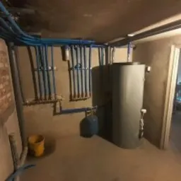 Piwnica z nową instalacją hydrauliczną: liczne niebieskie rury prowadzone pod sufitem i wzdłuż ściany, szary zasobnik na wodę, fragment ściany z cegły, żółte wiadro na podłodze.