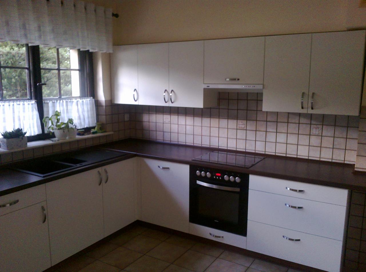 Jasna kuchnia z białymi szafkami i ciemnym blatem. Widoczny zlew, okno z firanką i płyta grzewcza. Prosta aranżacja w neutralnej kolorystyce.