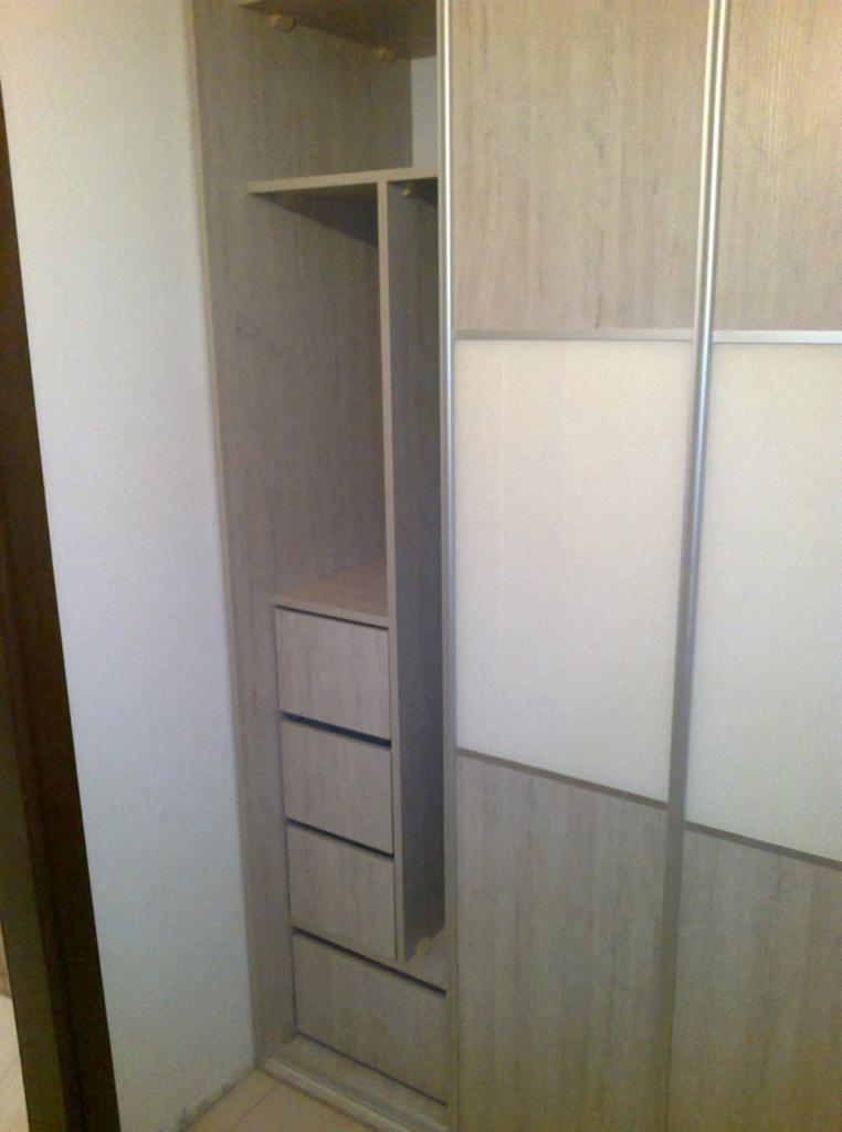 Jasna szafa wnękowa z przesuwnymi drzwiami, widoczne półki i szuflady. Minimalistyczny design, naturalne odcienie drewna i bieli.