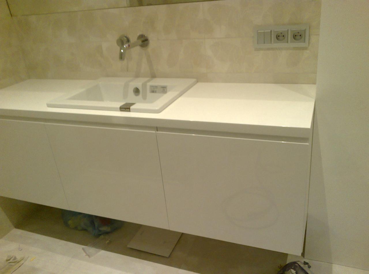 Biała szafka łazienkowa z prostokątną umywalką i chromowaną baterią na tle beżowych płytek. Widoczne gniazdka elektryczne. Minimalistyczny design.