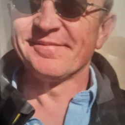 Portret mężczyzny w okularach przeciwsłonecznych i niebieskiej koszuli, z widocznym srebrnym łańcuszkiem na szyi, oświetlony z jednej strony, zarysowany ciemną kurtką.