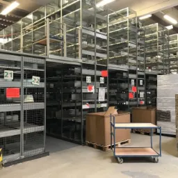 Wysokie, metalowe regały magazynowe z numeracją, częściowo wypełnione, z wózkiem transportowym i zapakowanymi paletami w tle, widok z dołu.