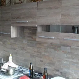 Kuchnia po remoncie: drewnopodobne szafki wiszące z metalowymi uchwytami nad blatem z kamienną mozaiką. Na blacie butelki i kuchenka z garnkiem.