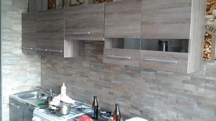 Kuchnia po remoncie: drewnopodobne szafki wiszące z metalowymi uchwytami nad blatem z kamienną mozaiką. Na blacie butelki i kuchenka z garnkiem.