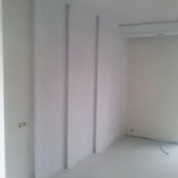 Szare wnętrze po remoncie, z widocznymi pionowymi wnękami na ścianie, gładkie ściany przygotowane do malowania. W rogu widoczny fragment instalacji.