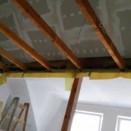 Skośny sufit poddasza z drewnianą konstrukcją i częściowo zamontowanymi płytami gipsowo-kartonowymi, widoczna drabina i okna w tle.