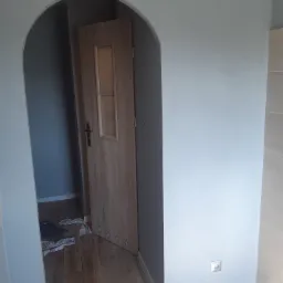 Szare wnętrze po odświeżeniu, z drewnianą podłogą, widoczne wejście łukowe i częściowo otwarte drzwi z przeszkleniem, na podłodze leżą szmaty.