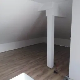 Układanie paneli podłogowych w przestrzeni poddasza z widoczną kolumną konstrukcyjną i narzędziami pracy.