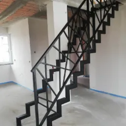 Metalowe schody o geometrycznym wzorze balustrady w surowym wnętrzu domu w trakcie wykończenia, widoczne zabezpieczenia folią malarską.