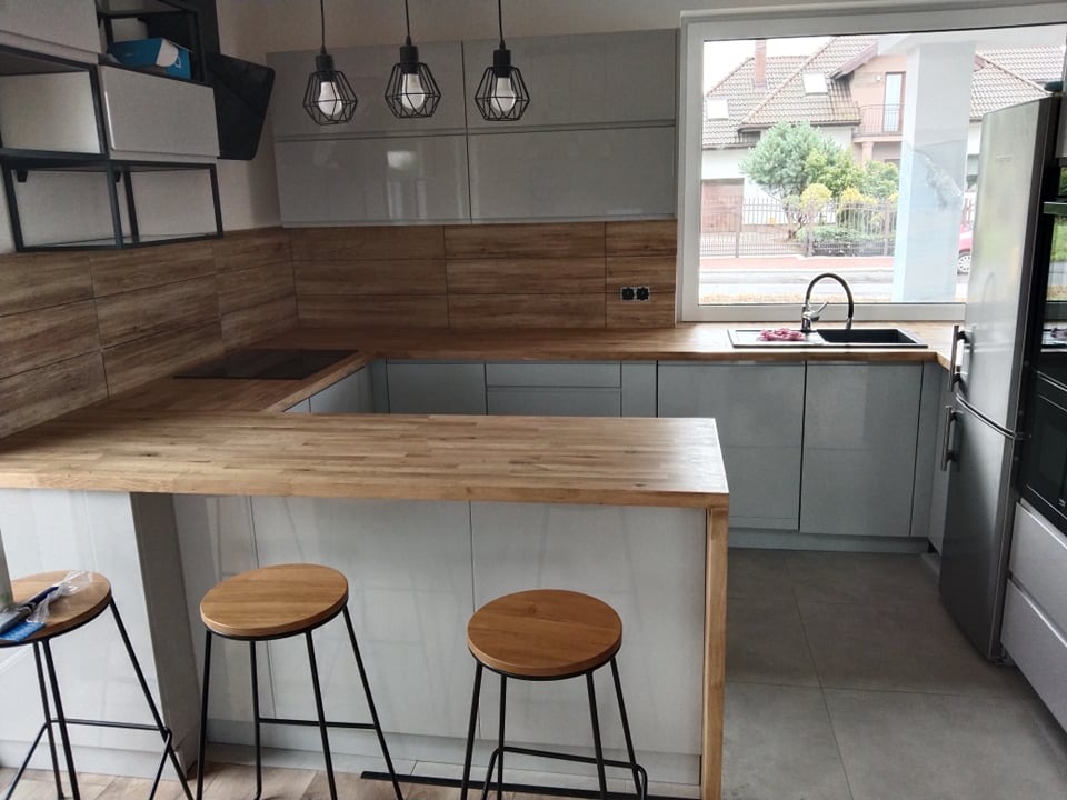 Nowoczesna kuchnia z szarymi szafkami, drewnianym blatem i barkiem z trzema hokerami. Nad blatem roboczym wiszą trzy lampy w metalowych kloszach. Widok na dom przez okno.