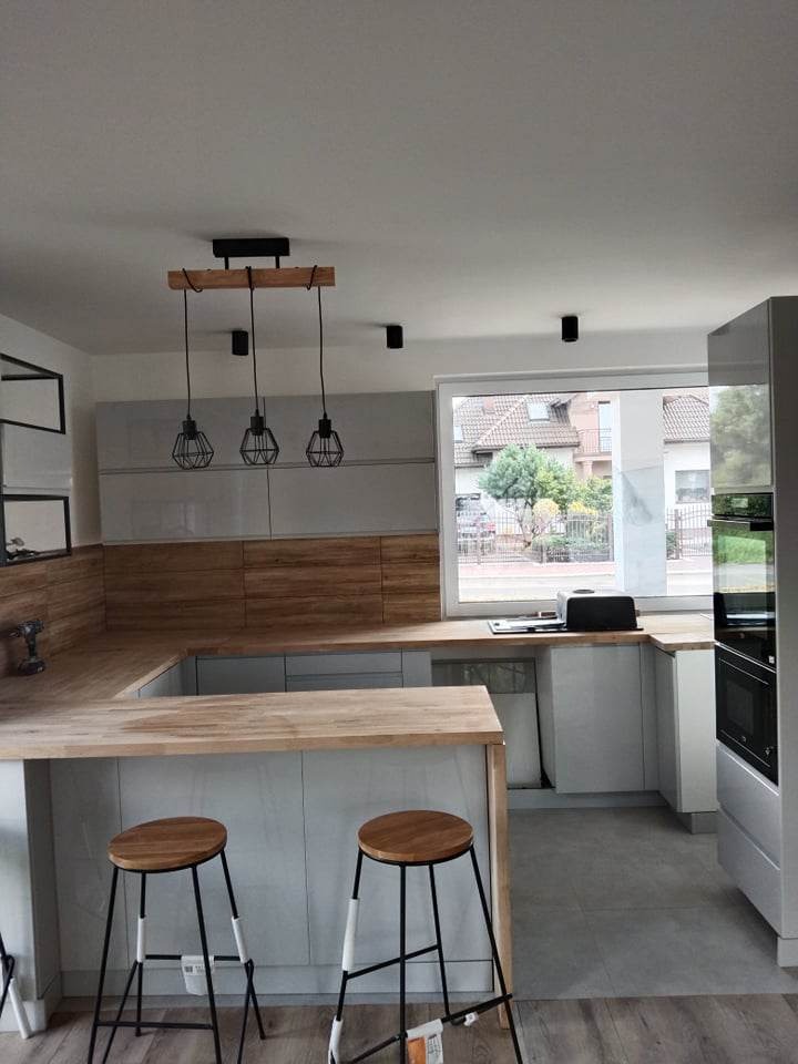 Nowoczesna kuchnia z drewnianym blatem, szarymi szafkami i metalowymi lampami wiszącymi, widok na osiedle domów przez okno.