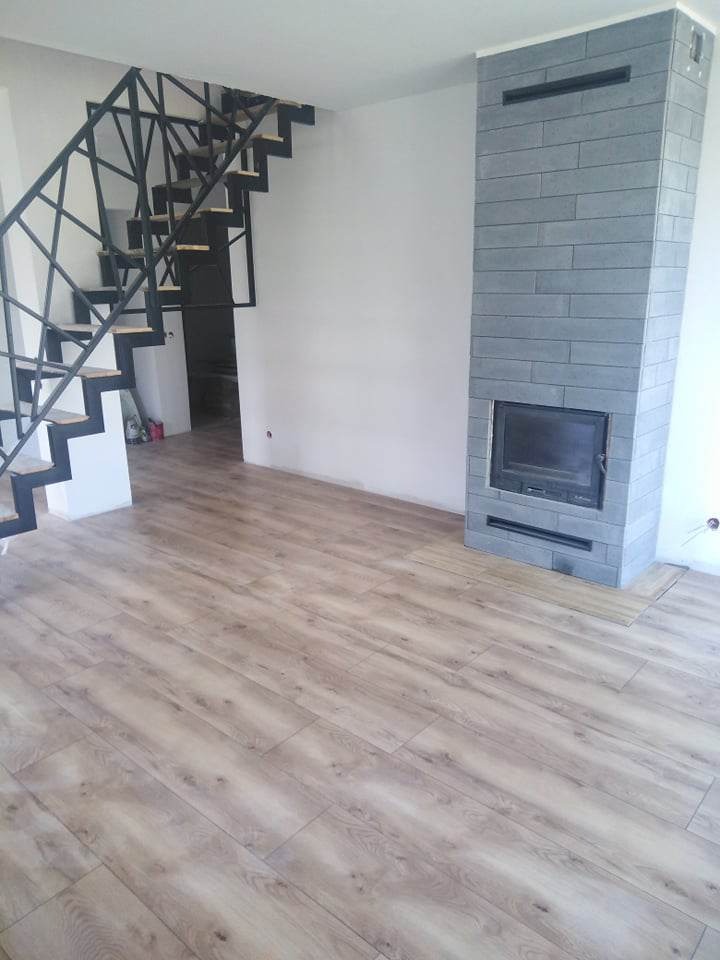 Salon po remoncie: świeżo ułożone panele podłogowe w odcieniach drewna, nowoczesne, metalowe schody z geometryczną balustradą oraz obłożony szarą cegłą kominek.