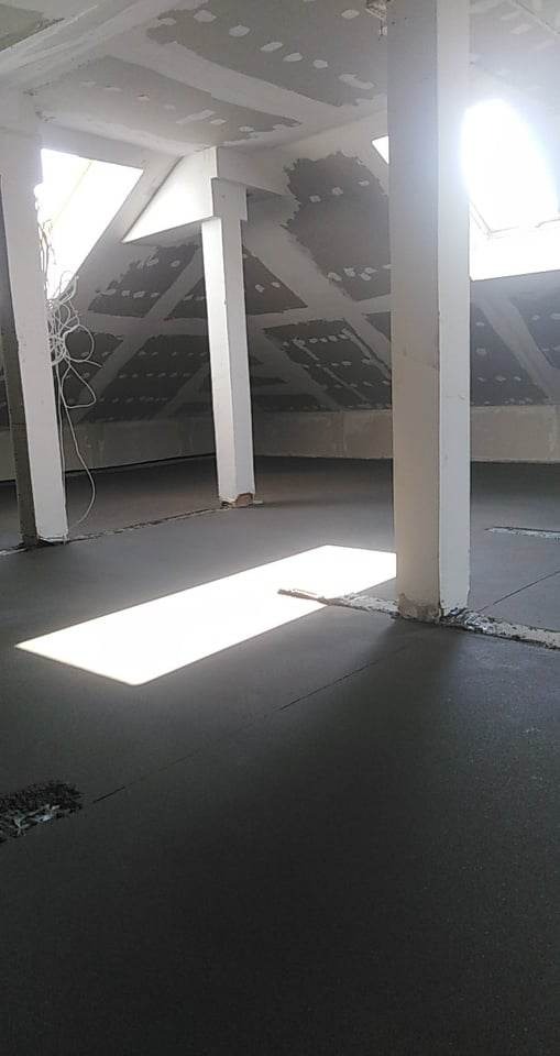 Poddasze w trakcie prac wykończeniowych: wylany betonowy podkład podłogowy, surowy sufit z widocznymi łączeniami płyt gipsowo-kartonowych, białe kolumny konstrukcyjne i wpadające przez okno dachowe...