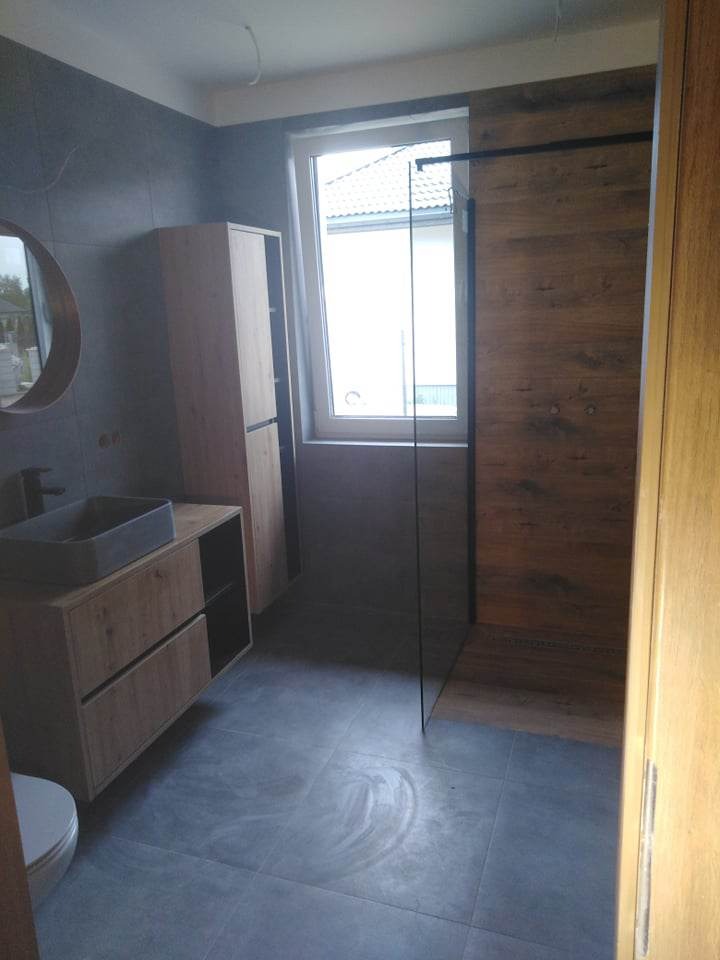Nowoczesna łazienka z szarymi płytkami podłogowymi i ściennymi, drewnianymi szafkami umywalkowymi z prostokątną umywalką, kabiną prysznicową typu walk-in z drewnianą ścianą i okrągłym lustrem.