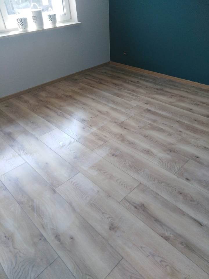 Jasne panele podłogowe ułożone w nowym pokoju z szarą i ciemnozieloną ścianą oraz trzema białymi kubkami na parapecie okiennym.