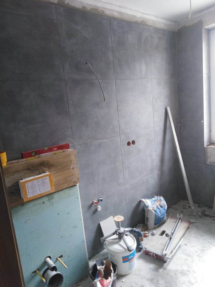 Szare płytki wielkoformatowe na ścianie w trakcie montażu, widoczne przygotowane otwory na instalacje, narzędzia glazurnicze i materiały budowlane, surowy stan wykończenia.