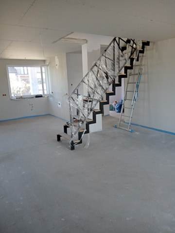 Nowoczesne, czarne schody z metalową balustradą, zabezpieczone folią ochronną, w surowym wnętrzu w trakcie remontu, z widoczną drabiną aluminiową w tle.