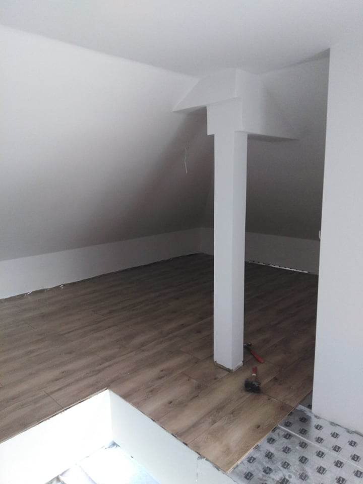 Układanie paneli podłogowych w przestrzeni poddasza z widoczną kolumną konstrukcyjną i narzędziami pracy.