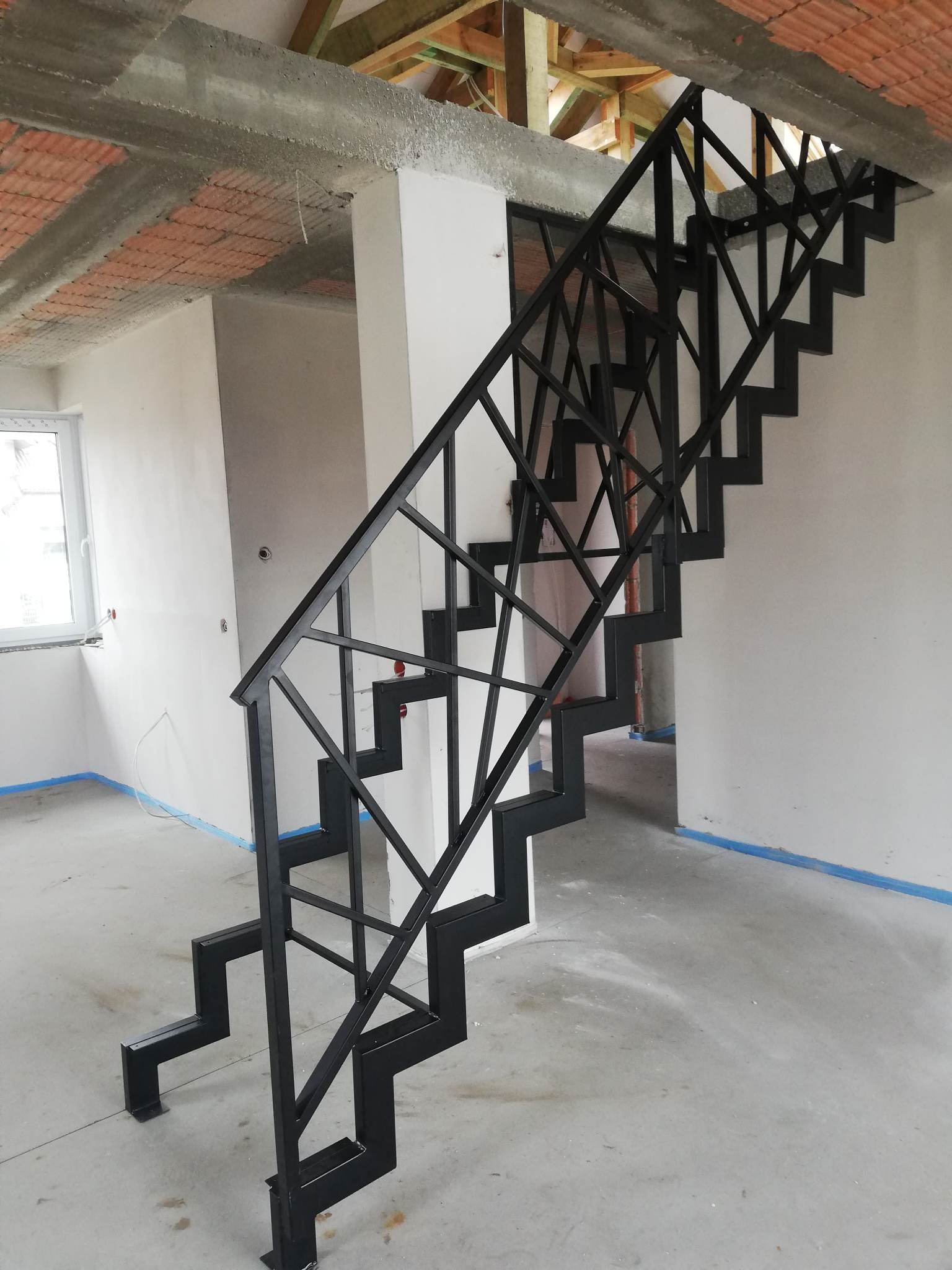 Metalowe schody o geometrycznym wzorze balustrady w surowym wnętrzu domu w trakcie wykończenia, widoczne zabezpieczenia folią malarską.
