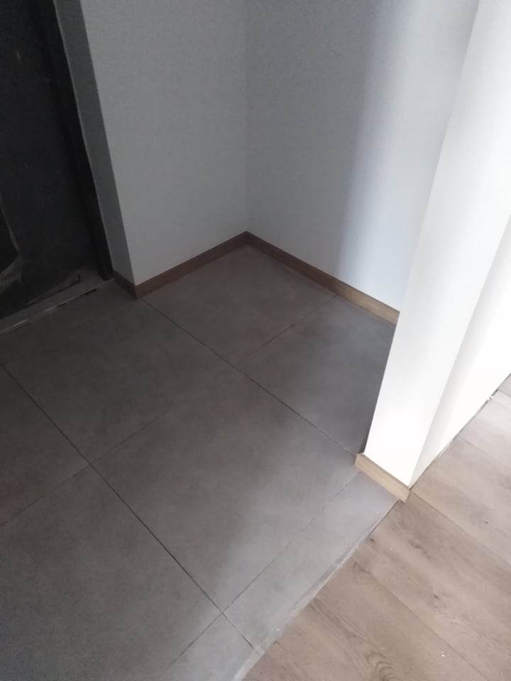 Szare płytki podłogowe w rogu pomieszczenia, z widoczną listwą przypodłogową i fragmentem drewnianej podłogi w tle, oświetlone naturalnym światłem.
