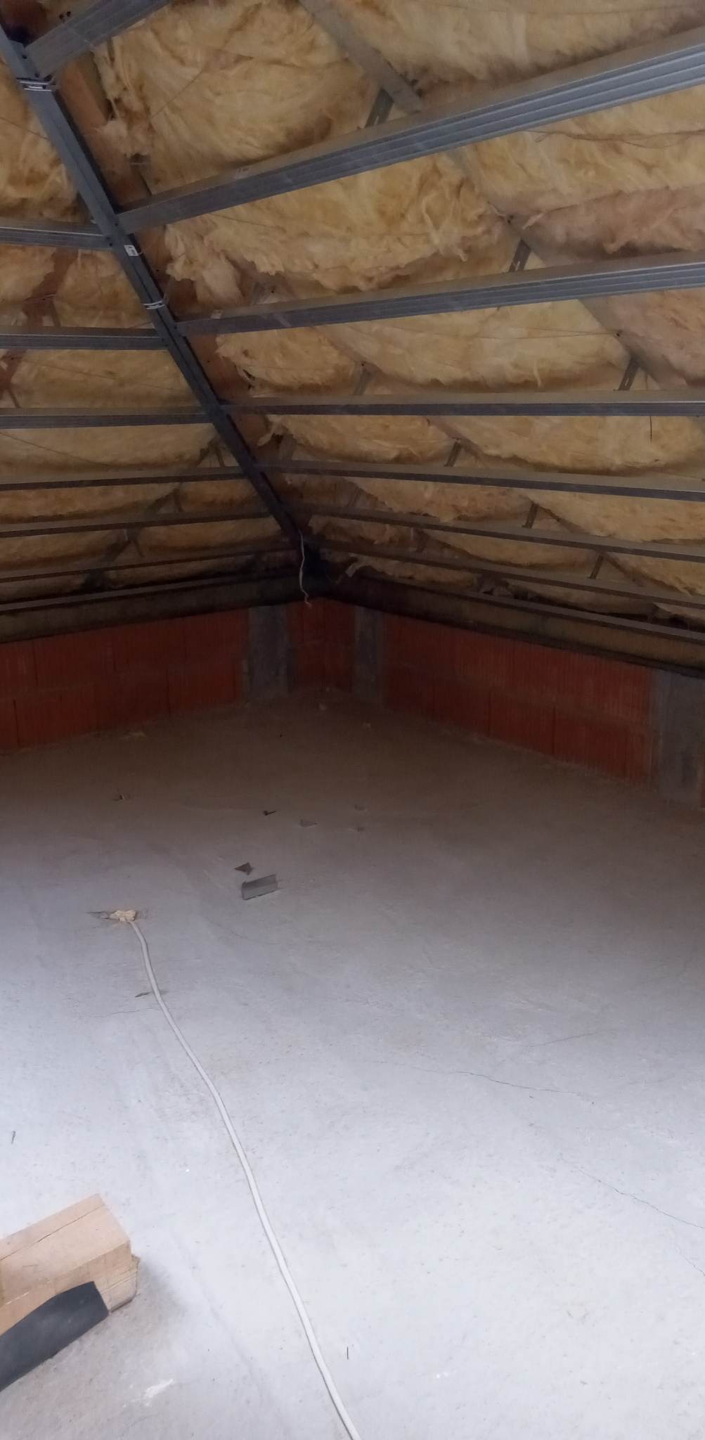 Poddasze w trakcie adaptacji: widoczne ocieplenie wełną mineralną pod metalową konstrukcją sufitu, ściany z czerwonej cegły oraz surowa betonowa podłoga z leżącym kablem elektrycznym.