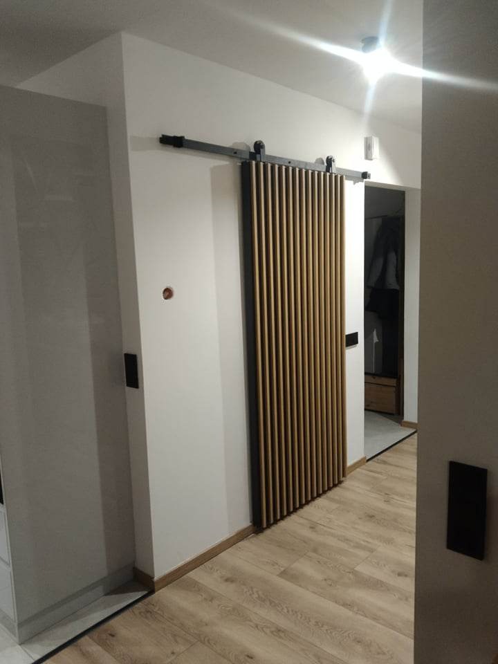 Nowoczesne drzwi przesuwne, wykonane z pionowych drewnianych listew w ciemnej ramie, zawieszone na czarnej szynie, zamontowane w jasnym wnętrzu z panelami podłogowymi.