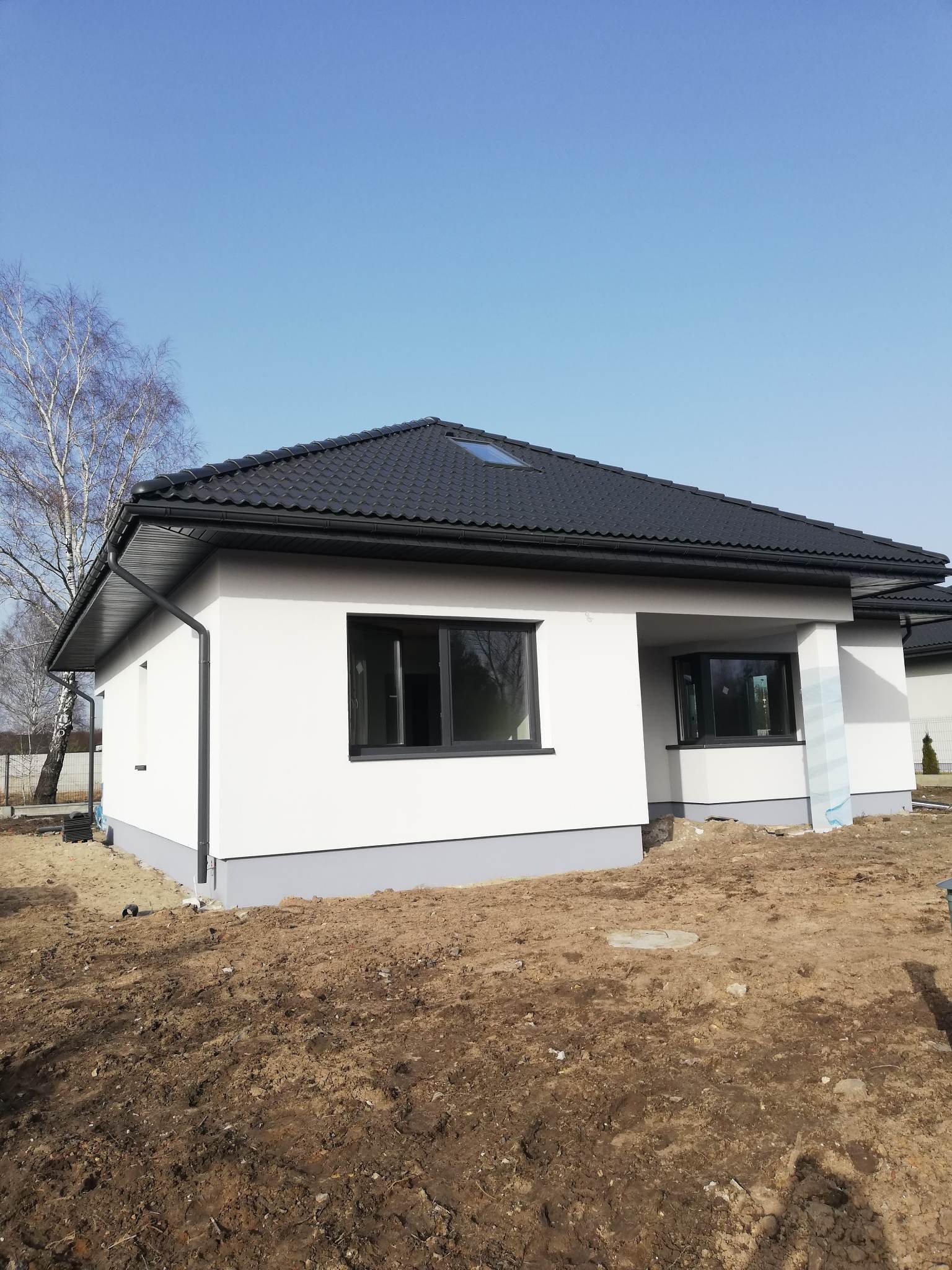 Biały, parterowy dom z czarnym dachem krytym dachówką, czarnymi oknami i szarą podmurówką, w trakcie prac wykończeniowych terenu wokół budynku.