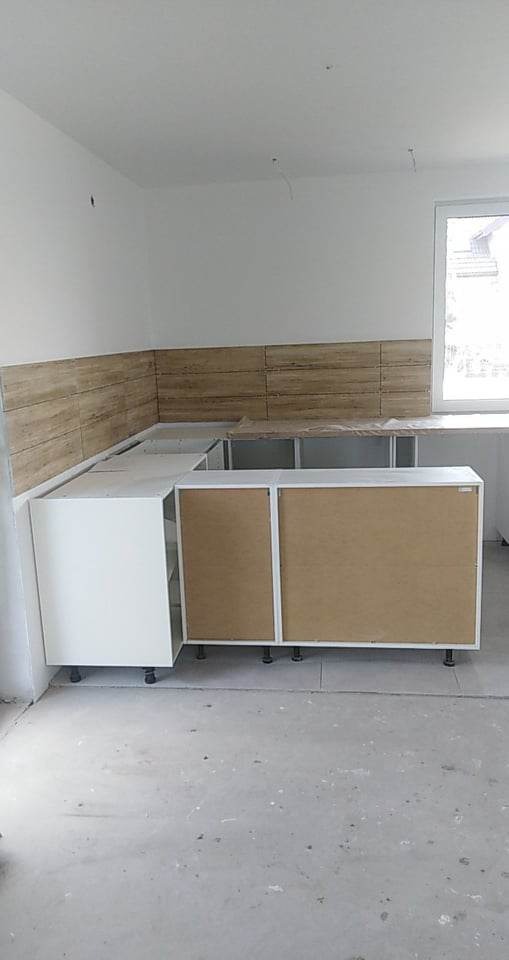 Częściowo zmontowane, białe szafki kuchenne z brązowymi frontami, ustawione pod ścianą z drewnopodobnymi płytkami, widoczne regulowane nóżki, surowy beton na podłodze, okno w tle.