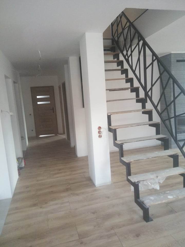 Wnętrze w trakcie wykańczania z drewnopodobną podłogą, białe ściany i schody z metalową balustradą o geometrycznym wzorze, zabezpieczone folią. Widoczny fragment kominka w tle.