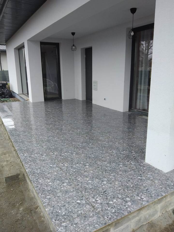 Zadaszony taras wyłożony szarymi płytkami imitującymi kamień, widoczne białe ściany budynku z czarnymi ramami okiennymi i drzwi oraz geometryczne lampy wiszące.