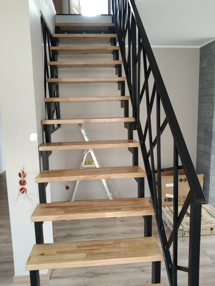 Nowoczesne schody z drewnianymi stopniami i czarną, geometryczną balustradą, częściowo zasłonięte przez rozłożoną drabinę i kartony w tle.