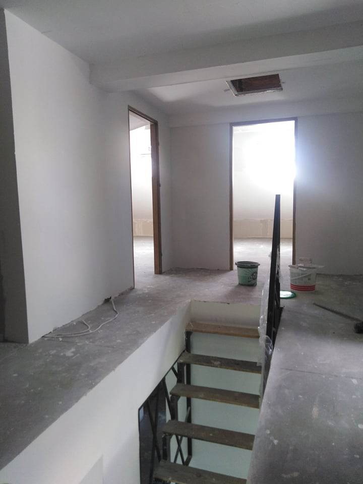 Wnętrze domu w trakcie prac wykończeniowych; świeżo pomalowane ściany na biało, widoczne schody z drewnianymi stopniami, dwa otwarte przejścia do innych pomieszczeń, puste wiaderka po farbie.