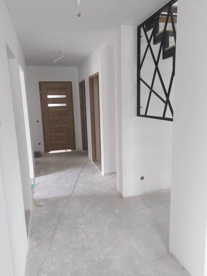 Korytarz w trakcie wykańczania: świeżo pomalowane białe ściany, surowa betonowa podłoga, drewniane drzwi wewnętrzne z poziomymi przeszkleniami i dekoracyjna, metalowa balustrada schodowa...