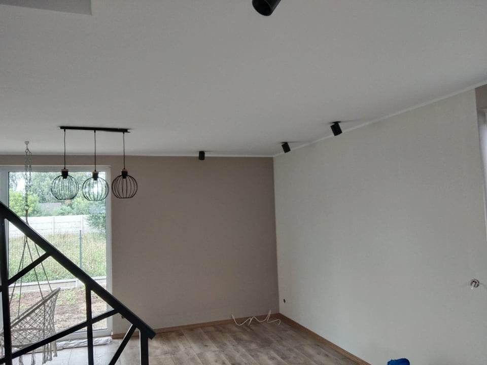 Świeżo pomalowane wnętrze z drewnianą podłogą, nowoczesnym oświetleniem sufitowym i wiszącymi lampami kulistymi, widoczne schody z czarną balustradą i okno z widokiem na zewnątrz.