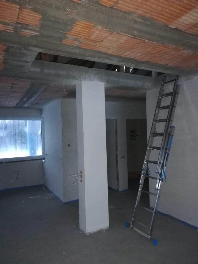 Wnętrze w trakcie prac wykończeniowych: surowy sufit z cegieł, otynkowane ściany, betonowa podłoga, aluminiowa drabina oparta o ścianę i biała kolumna na środku pomieszczenia.