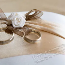 Dwa obrączki ślubne z wygrawerowanymi imionami i datą 16.04 na poduszce zdobionej kremową różą i brązowymi wstążkami.