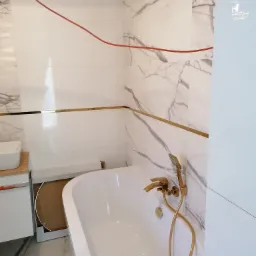 Biała wanna z złotą armaturą i marmurowe płytki na ścianach z poziomymi złotymi listwami, widoczny fragment szafki pod umywalkę i czerwony kabel na górze, w trakcie prac wykończeniowych.