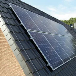 Panele fotowoltaiczne zamontowane na ciemnym, spadzistym dachu domu z cegły, widoczne okno i komin w tle drzewa.